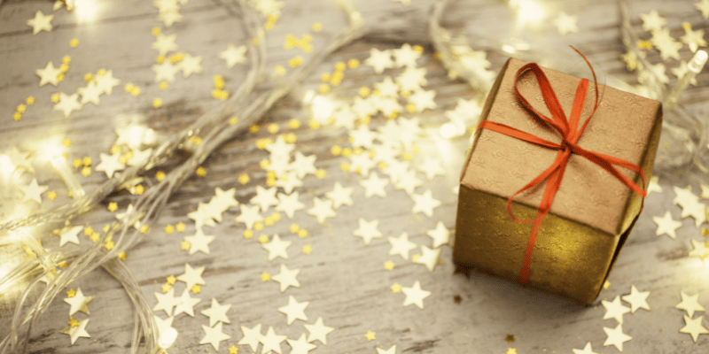 A table sprinkled with small golden star confetti, topped with warm fairy lights and a small brown paper wrapped present, with a red ribbon.
