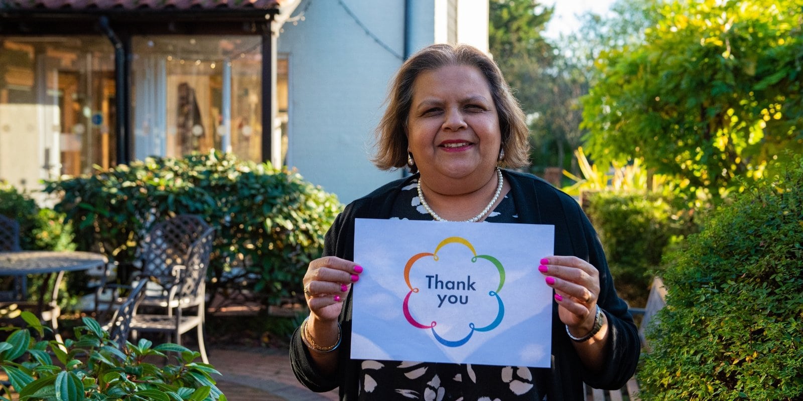 St Clare Hospice Head of Patient and Family Support Services, Sushma, is pictured smiling at the camera in the Hospice gardens whilst holding a sign that reads "Thank you".