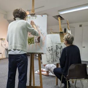 Two people can be seen in front of an easel drawing. One is standing up with the other sat in a chair.