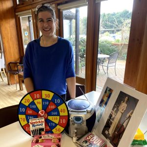 A woman stands behind a table with various items on display including a dart board, mini stereo, pictures 