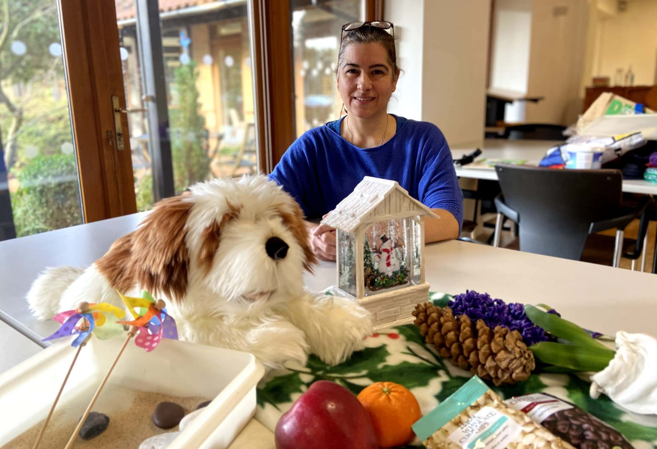 A woman sits behind a desk with an assortment of items including a large cuddly toy, a pine cone, a sand box with two pin wheels and a bird box
