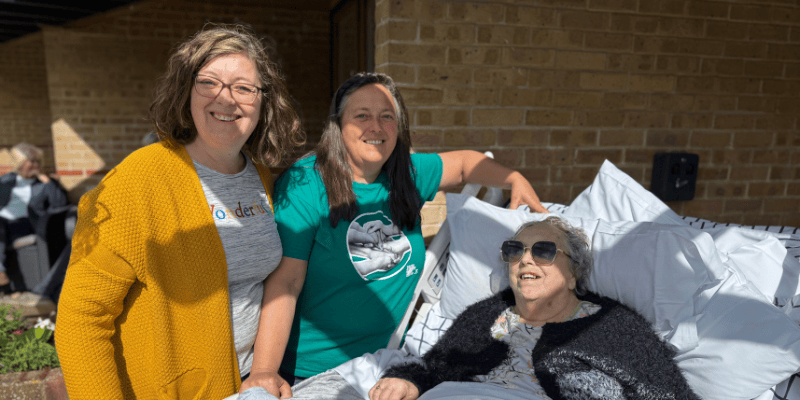 Photo of Wyn wearing sunglasses while in her Inpatient Unit bed pictured with her two daughters. All smile.
