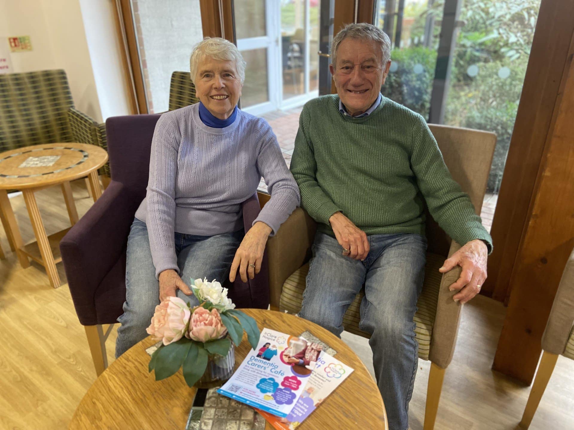 A husband and wife sit together behind a small coffee table. On the table is a small posy of flowers and some leaflets.