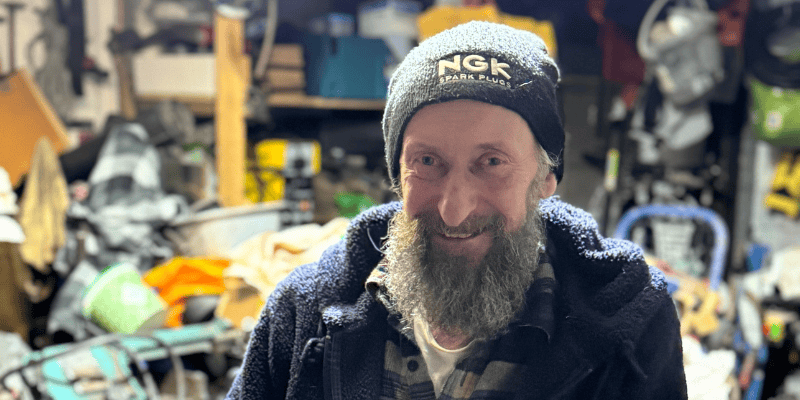 Nigel is pictured smiling wearing a beanie in front of his shed of tools and lawnmowers.