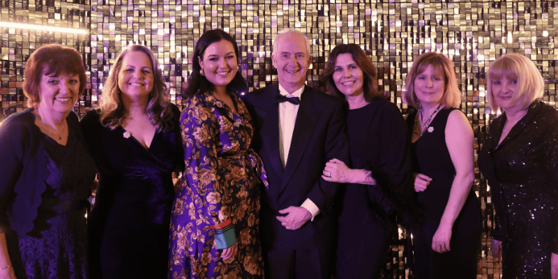 A photo of a group of women and a man can be seen. They all wear black-tie outfits and smile at the camera.