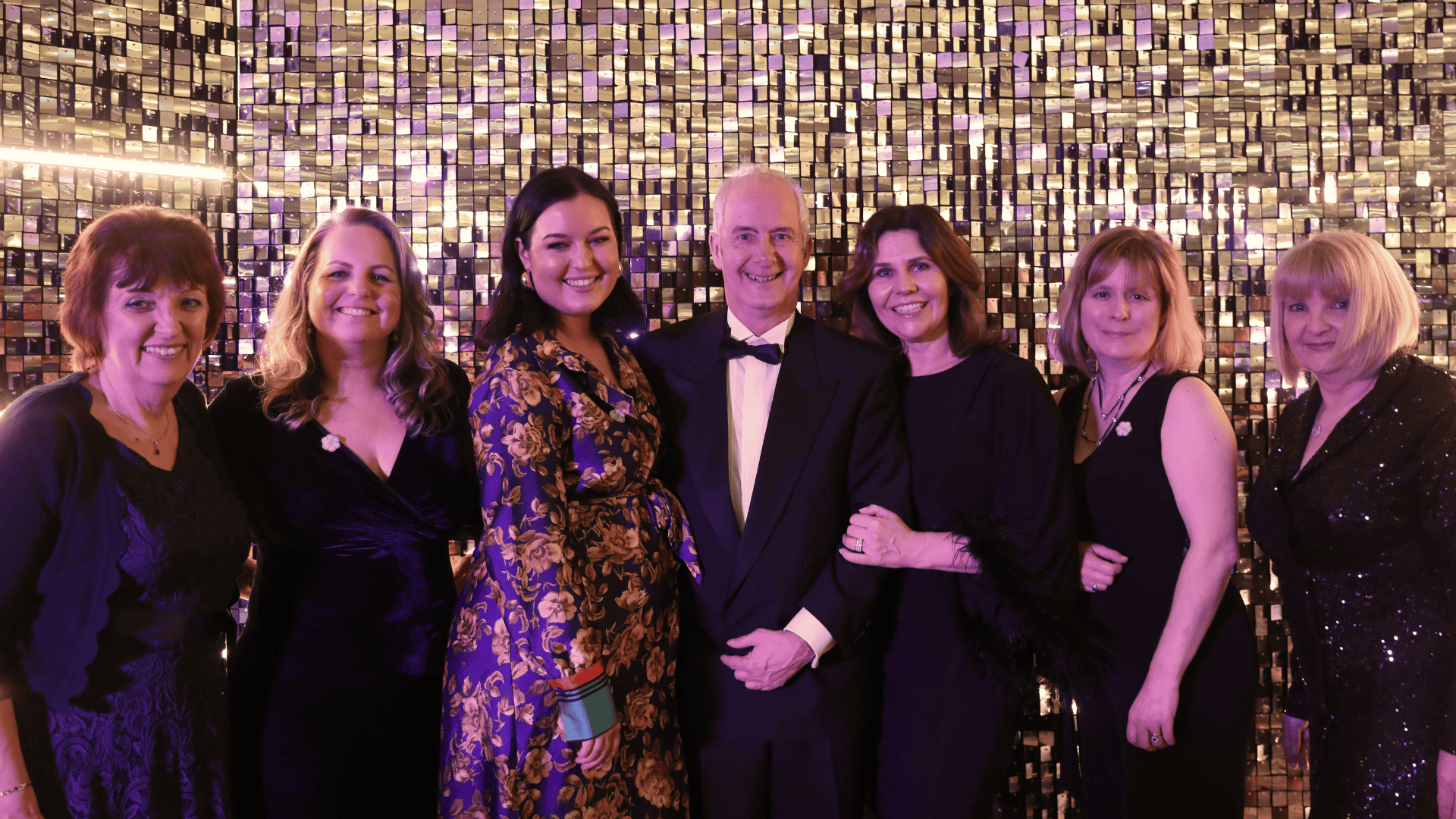 A photo of a group of women and a man can be seen. They all wear black-tie outfits and smile at the camera.